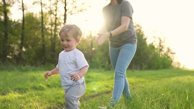 Toddler walking with parent in grass sunlit field where child smiles while adult supports steps family enjoying outdoor nature play near tree line warm light enhancing happy bond and gentle movement