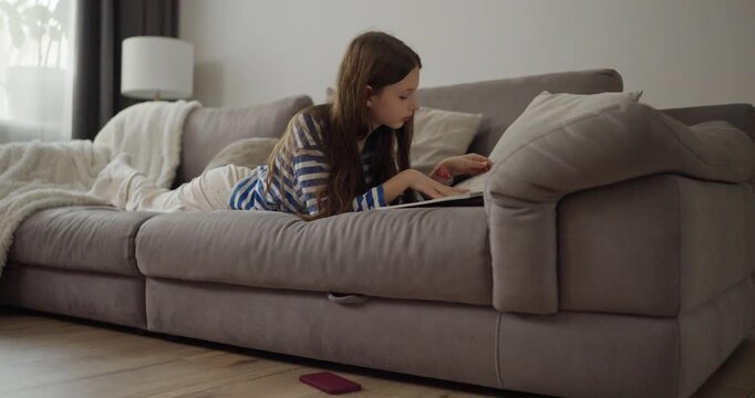 Teenage girl in striped sweater lying on cozy sofa and flipping through magazine at home during daytime. Quality rest and self-development at home