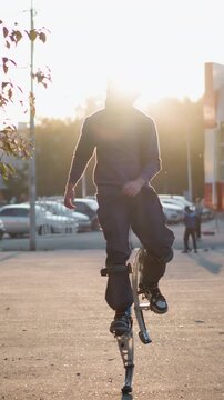 Front shot of man wearing gray hoodie and dark gray pants jumping on spring stilts on paved road in residential area surrounded by houses during sunset, with serious expression