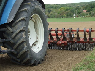 The rear wheel of a tractor is visible, a harrow is attached to the tractor