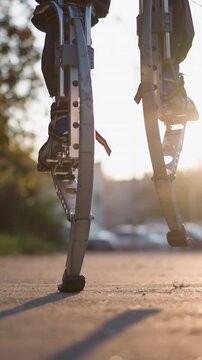 Close-up view of legs walking on spring stilts in suburban outdoor setting with dark pants and sneakers. Background features blurred trees, buildings, and a street. Low camera angle highlights