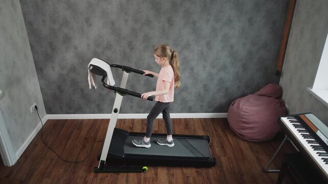 Top down view of teenage girl in pink shirt and patterned leggings walking on treadmill holding handles in home workout room with textured grey wall wooden floor partial piano and beanbag visible