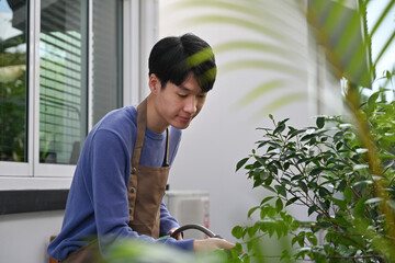 A Young Asian man in apron watering lush green plants outdoors, enjoying peaceful gardening near a...