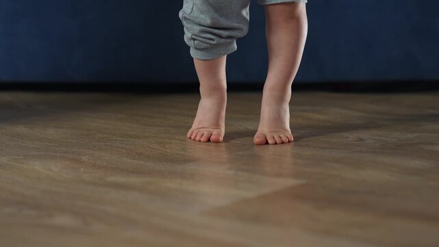 Toddler taking first tentative barefoot steps on hardwood floor showing foot movement and balance during walking practice with baby energy step by step development and playful exploration of new skill