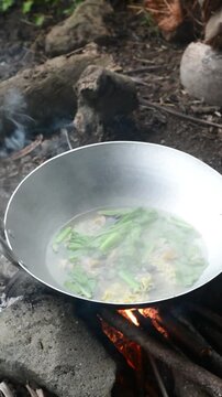 Indonesian woman preparing seblak over charcoal fire in a bamboo saung near rice fields. Traditional cooking method and countryside atmosphere.