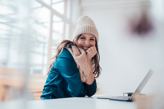 Happy businesswoman wearing knit hat sitting in front of laptop at home office