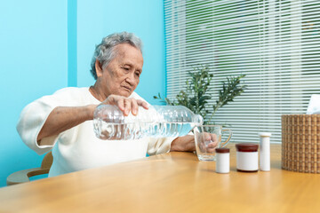 An elderly Asian woman pours drinking water from a plastic bottle into a glass to prepare to take...