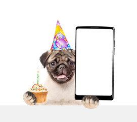 Happy Pug puppy wearing party cap holds capcake with candle and big smartphone with white blank screen in it paw, showing close to camera above white banner. Empty free space for mock up, banner