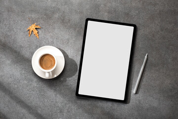 A mockup tablet with a blank screen, stylus and cup of coffee with autumn leaves on a gray stone desktop with sunlight from the window