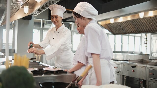 Female chef demonstrates to intern student girls, cooking class in catering restaurant kitchen, professional gourmet master in delicious cuisine, ingredients preparation in eatery, and culinary meals.