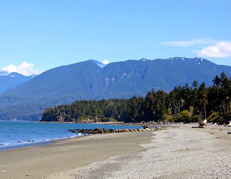 Sandy beach, rocky shoreline, evergreen forest, and distant mountains under a clear blue sky