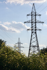Two Steel Lattice High Voltage Electricity Pylons (Transmission Towers) Standing Among Green Trees Against A Blue Sky With Clouds. Vertical.