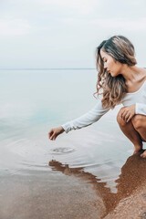 Asian woman squatting barefoot at lake edge creating water ripples with fingers, gazing at water...