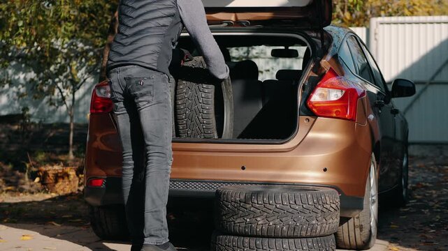 A man in jeans and a vest is loading car tires into the open trunk of a brown vehicle. Several stacked winter tires with studs are placed near the car, suggesting seasonal tire replacement