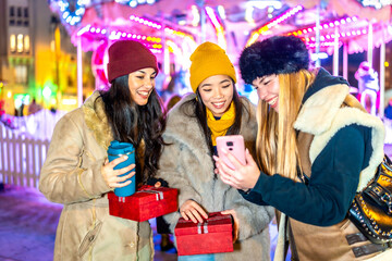 Happy friends using phone and holding christmas gifts at night with carousel lights in background