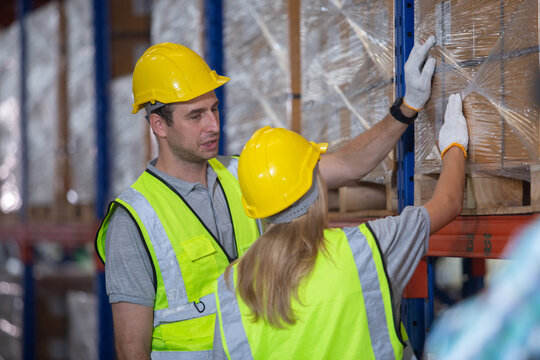 Male and female warehouse workers in safety gear inspecting packages on industrial shelves. Team coordination and inventory control in logistics operations.