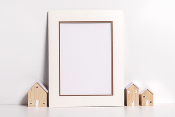 Empty white picture frame on a white wooden table with a minimalist small house.