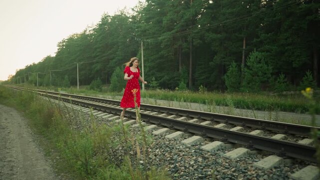 Side view of young woman in red dress running cautiously on rail sleeper through countryside area, surrounded by gravel, green vegetation, tall trees, and utility pole under soft natural daylight