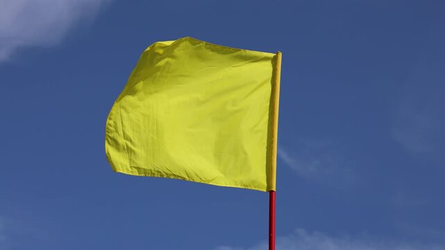 A bright yellow flag waves on a red pole against a clear blue sky, symbolizing caution or marking an area with bold visibility