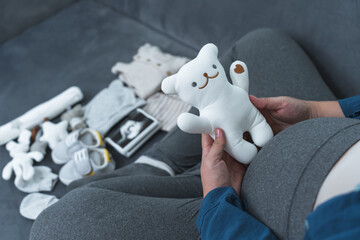 A pregnant woman sitting on a sofa holding small bear doll for her unborn baby. Baby clothes,...