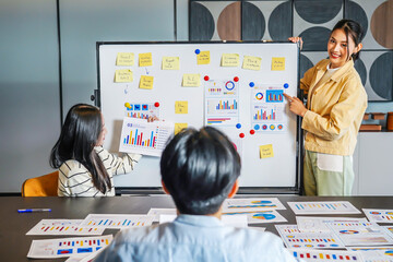 Young asian businesswoman conference speaker passionately explains business project strategy using a whiteboard, engaging a diverse team in collaborative brainstorming within a contemporary office