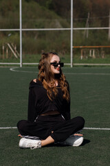 A girl in sportswear is sitting on a football field. She has dark glasses.