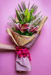 A woman's hand holding a beautiful bouquet of flowers on a pink background. A beautiful bouquet of flowers. Pink lilies, palm leaf, baby’s breath