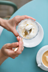 Overhead view of a woman enjoying vanilla ice cream in a glass bowl on the summer terrace, with a cappuccino nearby.