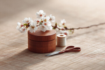 Fototapeta premium Elegant still life with a wooden box adorned with cherry blossoms, spool of thread, and scissors on a woven mat. Captures serenity, craft, and springtime beauty.