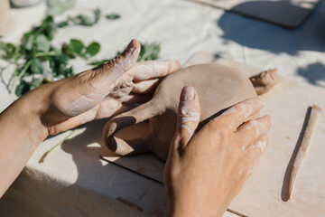 Hands shape a piece of clay into a distinct form in a spacious studio, surrounded by tools and greenery, showcasing the artistry of pottery making during a workshop