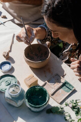 A person carefully paints delicate patterns on a clay cup at a pottery workshop held outdoors, surrounded by various tools and materials for ceramics