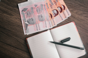 Cambodian Riel banknotes arranged on a wooden desk, next to an open notebook and pen. This image...