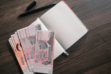 Cambodian Riel banknotes arranged on a wooden desk, next to an open notebook and pen. This image...