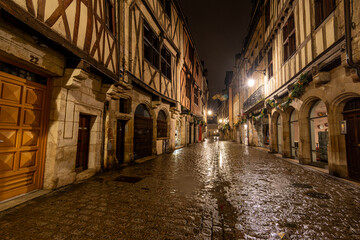 Night view of a wet medieval cobbled street in Dijon, France. The photograph highlights reflections from street lamps on the slick stones, historic architecture, and the atmospheric, moody ambiance.