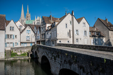 View of a bridge over the Eure River leading into the old town of Chartres, France, with Chartres Cathedral visible in the background under a clear blue sky. 