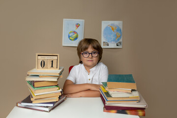 A boy in glasses sits at a desk against the background of geographical maps among books and a...