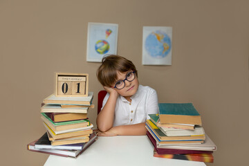 Tired boy in glasses sits at a desk on a background of geographical maps among books and a calendar...