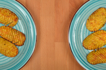 A high-angle shot showing two blue ceramic plates with baked and seasoned hasselback potatoes, placed on a light wooden background
