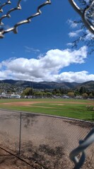 Baseball field seen through chain-link, with mountains in the background under a cloudy blue sky