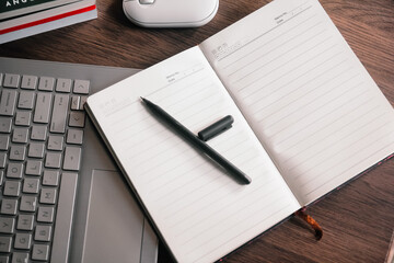 A pen and open notebook on a wooden desk, with a wireless mouse and laptop in the background. This...