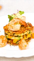 A layered dish of golden-brown fritters, topped with succulent shrimp, vibrant avocado, and mango, presented on a white plate.