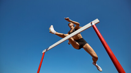 Female hurdler in low angle action leap against clear sky. Concept of overcoming barriers, strength, and determination in professional athletics training and competition.