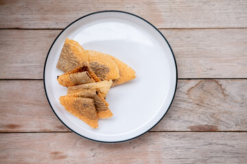  A small pile of folded Indonesian Kue Semprong cookies sits on a minimalist white plate, centered on a rustic wooden table, viewed from above.