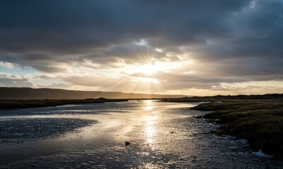 Sunlight breaks through dramatic clouds, casting rays over a wide, reflective river winding through a natural, grassy landscape at sunset or sunrise.