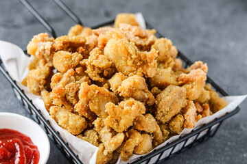 Golden-brown popcorn chicken or bite-sized fried chicken pieces, piled high in a black wire serving basket lined with white paper. A bowl of red ketchup is visible in the background.