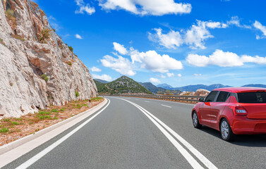 A red car driving on a scenic mountain highway with cliffs, blue sky, and clouds, symbolizing travel, adventure, and freedom on the open road.