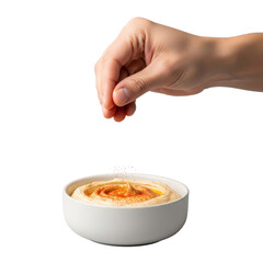 Light-skinned hand delicately sprinkling vibrant red paprika onto creamy beige hummus in a white ceramic bowl, isolated on a transparent background with soft studio light, concept of professional food