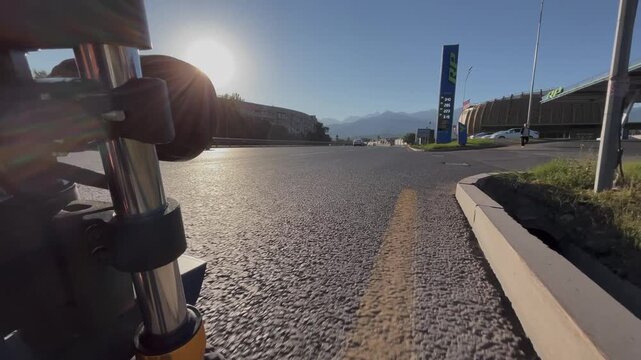 Camera mounted on moving transport captures drive along city road. Urban traffic scene with cars, asphalt, and street infrastructure. In the distance, mountain range rises above the city, combining ur