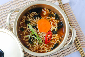 Top view of Korean Ramyeon (or Ramyun) in a gold-colored pot. The broth is filled with instant noodles, seaweed, and an egg yolk that's still runny. Chili slice and green scallions complete this dish.