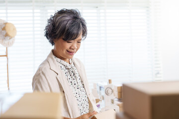 Senior asian woman fashion designer working in small family business packing product box for e commerce order shipping surrounded by fabric sewing machine and cardboard packages in bright studio